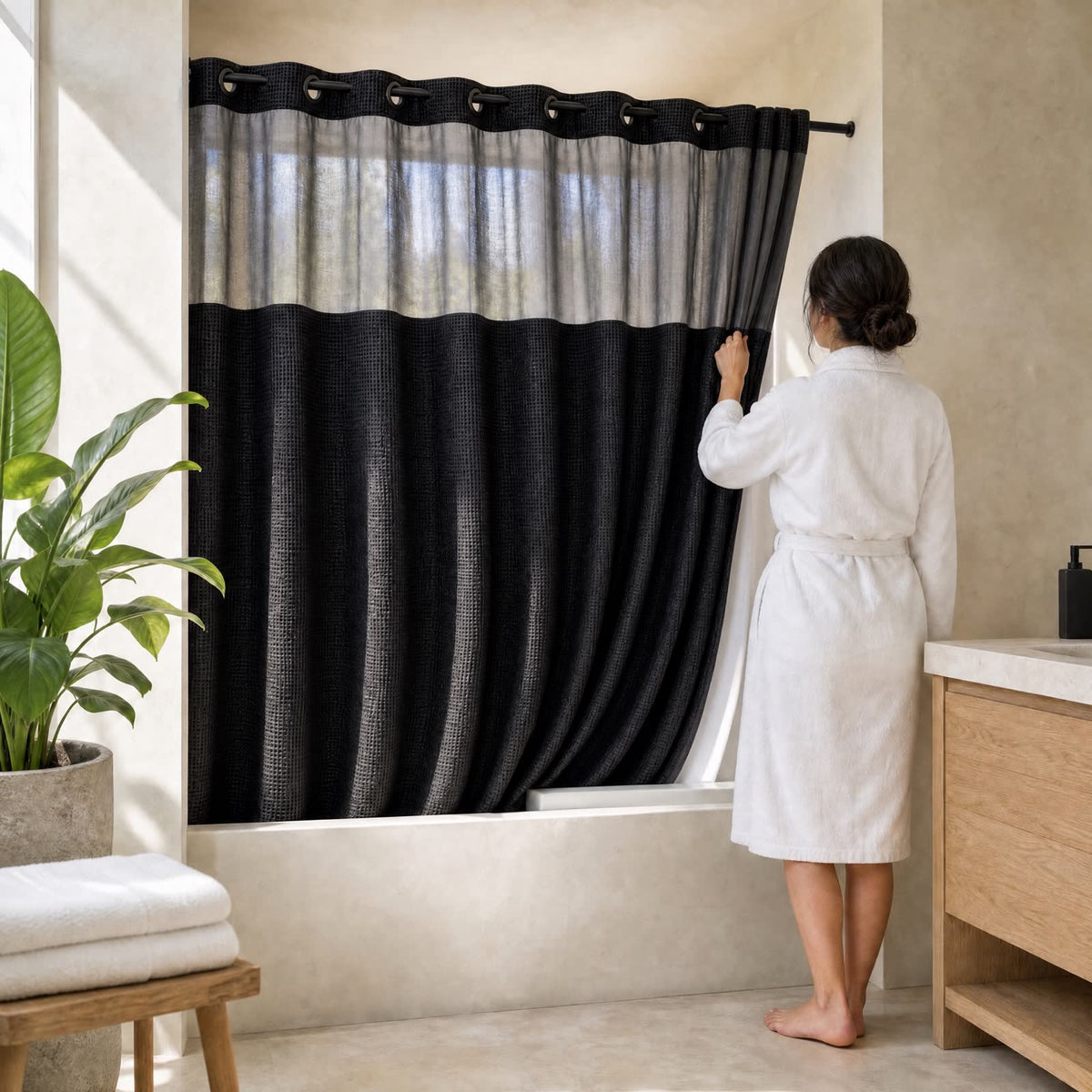 Une femme en peignoir tire le rideau de douche Aqualis™ noir en tissu gaufré dans une salle de bain lumineuse, la lumière traverse le voile fumé